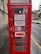 A traditional red telephone booth with signs indicating 'FOR SALE TO LET' along with contact details for inquiries. The booth is positioned on a rainy street, next to a stone building, with vehicles parked in the background.