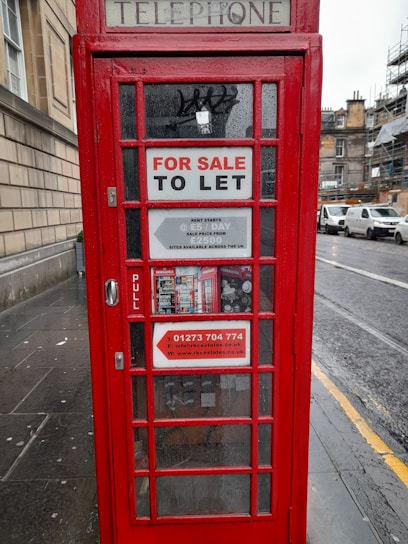 A traditional red telephone booth with signs indicating 'FOR SALE TO LET' along with contact details for inquiries. The booth is positioned on a rainy street, next to a stone building, with vehicles parked in the background.