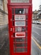 A traditional red telephone booth with signs indicating 'FOR SALE TO LET' along with contact details for inquiries. The booth is positioned on a rainy street, next to a stone building, with vehicles parked in the background.