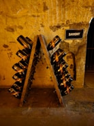 A rustic, dimly-lit wine cellar with an A-frame riddling rack holding numerous champagne bottles tilted downwards. The stone walls have a textured and aged appearance, with a small sign labeled 'SAVIGNY' mounted on the wall. The setting suggests an authentic and historic ambiance.