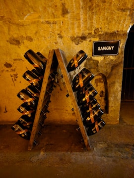 A rustic, dimly-lit wine cellar with an A-frame riddling rack holding numerous champagne bottles tilted downwards. The stone walls have a textured and aged appearance, with a small sign labeled 'SAVIGNY' mounted on the wall. The setting suggests an authentic and historic ambiance.
