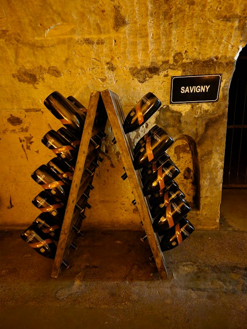A rustic, dimly-lit wine cellar with an A-frame riddling rack holding numerous champagne bottles tilted downwards. The stone walls have a textured and aged appearance, with a small sign labeled 'SAVIGNY' mounted on the wall. The setting suggests an authentic and historic ambiance.