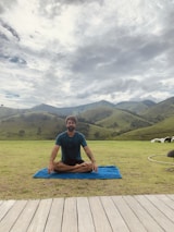A person is seated cross-legged on a blue mat in an open grassy area, surrounded by rolling green hills under a cloudy sky. Wooden planks form a path in the foreground, and the peaceful setting suggests a moment of meditation or relaxation in nature.