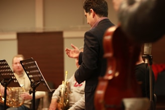A conductor stands with his back to the camera, possibly leading an orchestra or ensemble. Brass musicians are seated with music stands in front of them, and a string instrument's body is visible in the foreground. The setting appears to be a rehearsal or practice space.