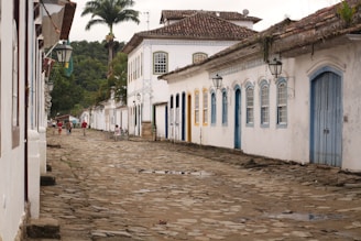 A narrow cobblestone street lined with white colonial-style buildings featuring colorful doors and windows. Large palm trees rise in the background, while a few people are walking along the street, some with bicycles. The scene conveys an old-world charm with its rustic architecture and serene atmosphere.