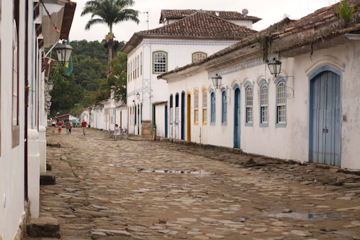 A narrow cobblestone street lined with white colonial-style buildings featuring colorful doors and windows. Large palm trees rise in the background, while a few people are walking along the street, some with bicycles. The scene conveys an old-world charm with its rustic architecture and serene atmosphere.