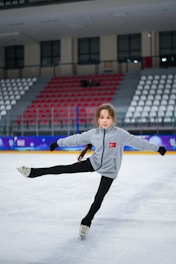 A young girl is ice skating in an indoor rink, wearing a gray jacket with a Turkish flag patch on it. Her arms and one leg are extended, suggesting she is performing a skating move. The background shows empty red and white spectator seats.