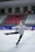 A young girl is ice skating in an indoor rink, wearing a gray jacket with a Turkish flag patch on it. Her arms and one leg are extended, suggesting she is performing a skating move. The background shows empty red and white spectator seats.