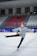 A young girl is ice skating in an indoor rink, wearing a gray jacket with a Turkish flag patch on it. Her arms and one leg are extended, suggesting she is performing a skating move. The background shows empty red and white spectator seats.