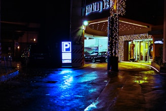 A parking area illuminated at night with vivid neon and warm yellow lights. A large blue parking sign stands out prominently. The entrance is decorated with string lights, and several cars are visible parked inside. Wet pavement reflects the colorful lights, adding a vibrant effect to the scene.