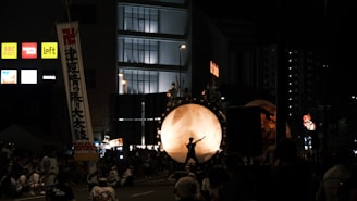 A nighttime festival scene with a large drum prominently displayed in the center, surrounded by performers. The drum is illuminated from behind, casting the silhouette of a drummer in action. Crowds of people are gathered around, some sitting and others standing, while banners and commercial signs from nearby buildings are visible.
