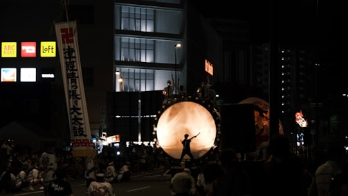 A nighttime festival scene with a large drum prominently displayed in the center, surrounded by performers. The drum is illuminated from behind, casting the silhouette of a drummer in action. Crowds of people are gathered around, some sitting and others standing, while banners and commercial signs from nearby buildings are visible.