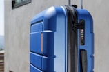 A close-up view of a large, hard-shell suitcase with a blue textured surface. The suitcase features a black combination lock and zipper on the side. The background consists of a neutral-colored wall and part of a window.