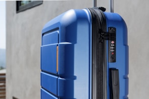A close-up view of a large, hard-shell suitcase with a blue textured surface. The suitcase features a black combination lock and zipper on the side. The background consists of a neutral-colored wall and part of a window.