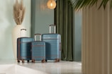 A set of three matching blue suitcases with brown accents is neatly arranged in a stylish interior space. The suitcases are positioned on a glossy, reflective floor, and a tall, white vase with pampas grass sits nearby. The background features a rich green curtain and a warm, woven pendant light hanging from the ceiling, creating a sophisticated and modern ambiance.