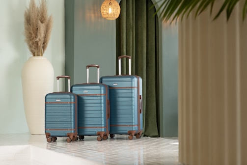 A trio of blue suitcases with brown accents is neatly arranged against a backdrop featuring green curtains. To the left, a large, white decorative vase is filled with pampas grass. The scene is set in a modern, well-lit space, featuring clean lines and a light-colored floor with a glossy finish.