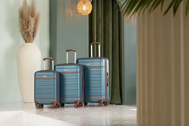 A trio of blue suitcases with brown accents is neatly arranged against a backdrop featuring green curtains. To the left, a large, white decorative vase is filled with pampas grass. The scene is set in a modern, well-lit space, featuring clean lines and a light-colored floor with a glossy finish.