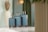 A trio of blue suitcases with brown accents is neatly arranged against a backdrop featuring green curtains. To the left, a large, white decorative vase is filled with pampas grass. The scene is set in a modern, well-lit space, featuring clean lines and a light-colored floor with a glossy finish.