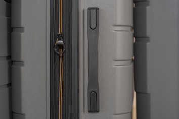 A close-up view of luggage surfaces tightly packed together, showing a combination of smooth and textured materials. There is a visible zipper with two thin yellow lines on one of the suitcases, and a prominent handle situated between them. The lighting highlights the subtle texture of the fabric.