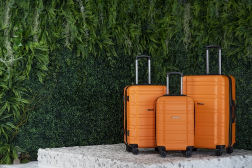 Three orange suitcases of varying sizes are neatly arranged on a textured platform. The background features lush, green foliage giving a natural and fresh vibe.