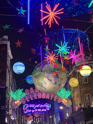 A vibrant street decoration with illuminated stars, planets, and neon signs. A large globe and colorful celestial elements are suspended, with words like 'Carnaby Celebrates' and 'Is this just fantasy?' visible amidst the lights.