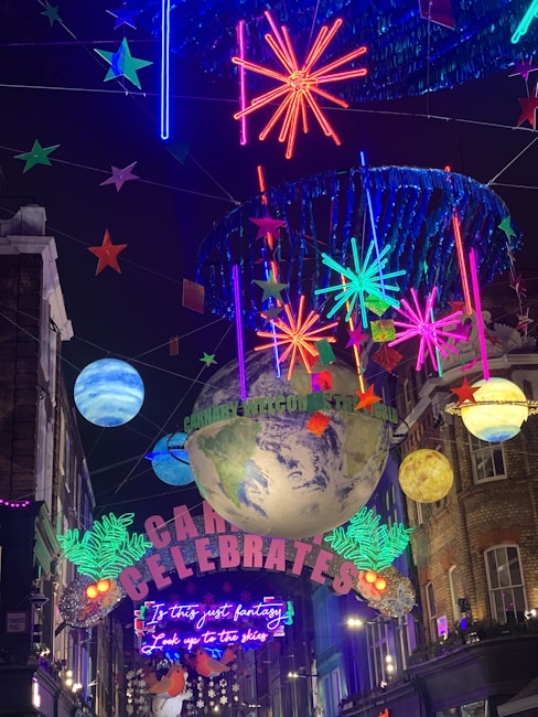 A vibrant street decoration with illuminated stars, planets, and neon signs. A large globe and colorful celestial elements are suspended, with words like 'Carnaby Celebrates' and 'Is this just fantasy?' visible amidst the lights.