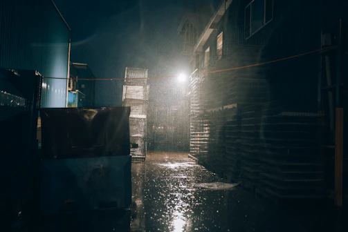 A rain-soaked industrial alley with flickering fluorescent lights casting eerie shadows on rusted pipes.