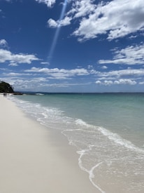 A tranquil beach scene with clear turquoise waters gently lapping onto a pristine white sandy shoreline. The sky is a vibrant blue, dotted with a few white, fluffy clouds. In the distance, a small stretch of greenery meets the ocean.