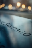 A close-up of a reflective metallic surface with the word 'SANTIAGO' engraved on it. In the background, there are warm, blurred lights creating a soft bokeh effect.