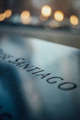 A close-up of a reflective metallic surface with the word 'SANTIAGO' engraved on it. In the background, there are warm, blurred lights creating a soft bokeh effect.