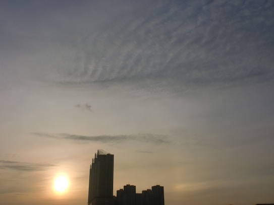 The image features a serene city skyline during sunset or sunrise. The sun is low on the horizon, emitting a soft glow, while clouds fill the sky creating a delicate pattern. A tall building and a few shorter buildings are visible in the foreground.