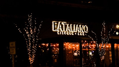 A warmly lit restaurant with a sign that reads 'Eataliano Kitchen & Bar'. The establishment is adorned with string lights on the exterior, creating an inviting and cozy atmosphere. Trees flanking the entrance are also wrapped in lights, enhancing the festive look. A 'No Parking Fire Lane' sign is visible on the left.