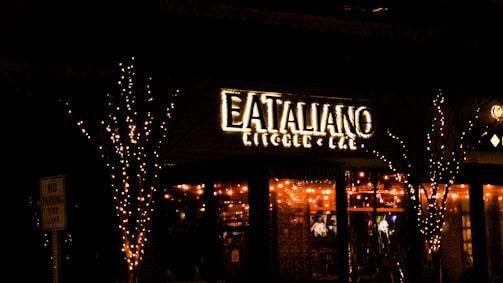 A warmly lit restaurant with a sign that reads 'Eataliano Kitchen & Bar'. The establishment is adorned with string lights on the exterior, creating an inviting and cozy atmosphere. Trees flanking the entrance are also wrapped in lights, enhancing the festive look. A 'No Parking Fire Lane' sign is visible on the left.
