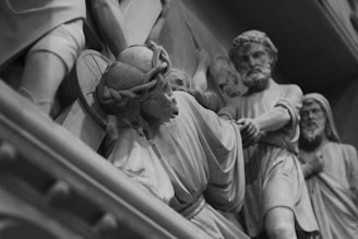 A detailed stone carving of several figures engaged in an emotional and dynamic scene. The central figure appears to be wearing a crown of thorns, suggesting a religious narrative. Other figures are depicted in flowing robes, with one figure grasping the arm of another, conveying tension and movement.