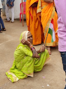 An elderly woman in a bright green sari sits on the ground holding a small Indian flag. She wears colorful bangles and a traditional headscarf. Nearby, other people are standing, some wearing orange saris, and there is a pink plastic chair in the background.