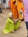 An elderly woman in a bright green sari sits on the ground holding a small Indian flag. She wears colorful bangles and a traditional headscarf. Nearby, other people are standing, some wearing orange saris, and there is a pink plastic chair in the background.