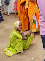 An elderly woman in a bright green sari sits on the ground holding a small Indian flag. She wears colorful bangles and a traditional headscarf. Nearby, other people are standing, some wearing orange saris, and there is a pink plastic chair in the background.