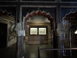 An ornately decorated and dimly lit room with intricate carvings and arches. The central area features a display of two framed artworks illuminated by focused lighting. The decor includes rich patterns on the ceiling and walls, adding to the historical ambiance of the space.