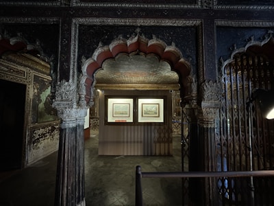 An ornately decorated and dimly lit room with intricate carvings and arches. The central area features a display of two framed artworks illuminated by focused lighting. The decor includes rich patterns on the ceiling and walls, adding to the historical ambiance of the space.