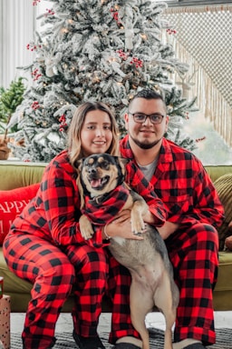 A couple wearing matching red and black plaid pajamas sits together on a couch, holding a dog. They are in front of a decorated Christmas tree with white snow-like effects and red ornaments. Presents are placed nearby, and the ambiance is cozy and festive.