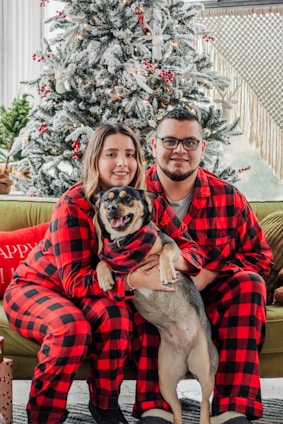 A couple wearing matching red and black plaid pajamas sits together on a couch, holding a dog. They are in front of a decorated Christmas tree with white snow-like effects and red ornaments. Presents are placed nearby, and the ambiance is cozy and festive.
