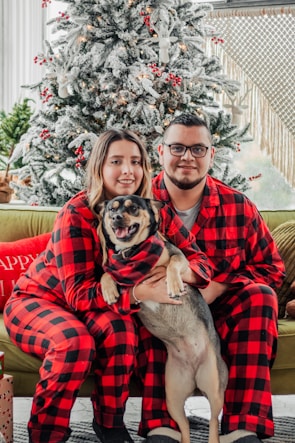A couple wearing matching red and black plaid pajamas sits together on a couch, holding a dog. They are in front of a decorated Christmas tree with white snow-like effects and red ornaments. Presents are placed nearby, and the ambiance is cozy and festive.