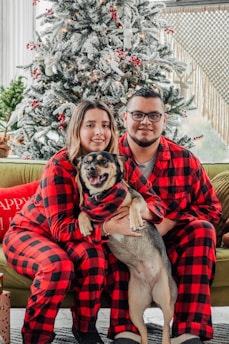 A couple wearing matching red and black plaid pajamas sits together on a couch, holding a dog. They are in front of a decorated Christmas tree with white snow-like effects and red ornaments. Presents are placed nearby, and the ambiance is cozy and festive.