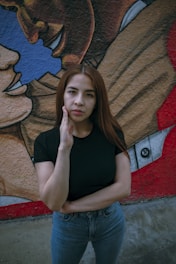 A woman with long brown hair, wearing a black t-shirt and blue jeans, stands in front of a colorful mural. The mural behind her depicts intertwined faces with blue, brown, and red colors, creating a vibrant backdrop.
