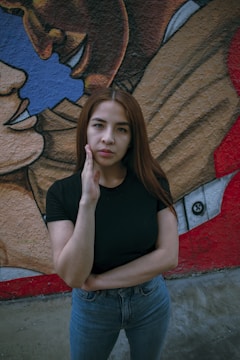 A woman with long brown hair, wearing a black t-shirt and blue jeans, stands in front of a colorful mural. The mural behind her depicts intertwined faces with blue, brown, and red colors, creating a vibrant backdrop.