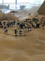 A detailed miniature scene built with LEGO blocks depicting an ancient desert setting. There are numerous LEGO figures, including camels and people, positioned around what seems to be a marketplace or village square. The background includes a pyramid structure, and various small constructs like tools or goods. The sand-colored base adds to the desert ambiance.