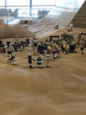 A detailed miniature scene built with LEGO blocks depicting an ancient desert setting. There are numerous LEGO figures, including camels and people, positioned around what seems to be a marketplace or village square. The background includes a pyramid structure, and various small constructs like tools or goods. The sand-colored base adds to the desert ambiance.