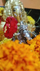 A small metallic statue of the Hindu deity Ganesha is surrounded by vibrant flowers, including red and yellow blossoms. The background features a blurred figure draped in gold fabric, enhancing the decorative and spiritual atmosphere.