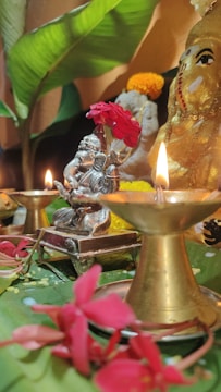 A traditional spiritual setup features a small statue holding bright red flowers and surrounded by lit brass oil lamps. The arrangement includes vibrant pink flowers and large green leaves, creating a serene and devotional atmosphere.