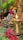 A traditional spiritual setup features a small statue holding bright red flowers and surrounded by lit brass oil lamps. The arrangement includes vibrant pink flowers and large green leaves, creating a serene and devotional atmosphere.